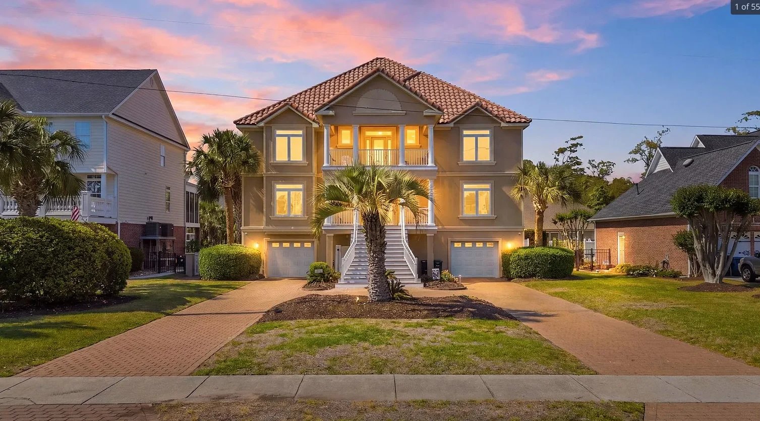 Waterfront house with two-story structure, lit windows, palm trees, manicured landscaping at dusk with pastel sky